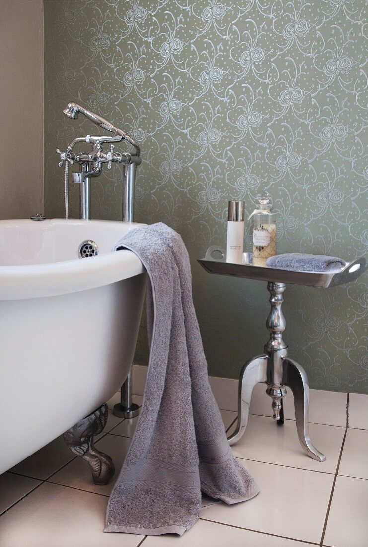Vintage Badewanne mit Löwenfüssen neben postmodernem, versilberten Beistelltischchen, an Wand gemusterte Flüssigtapete