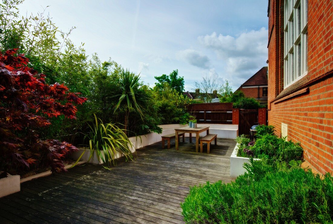 Bushes and palm trees in raised beds surrounding wooden terrace with table and benches adjoining brick facade of converted church