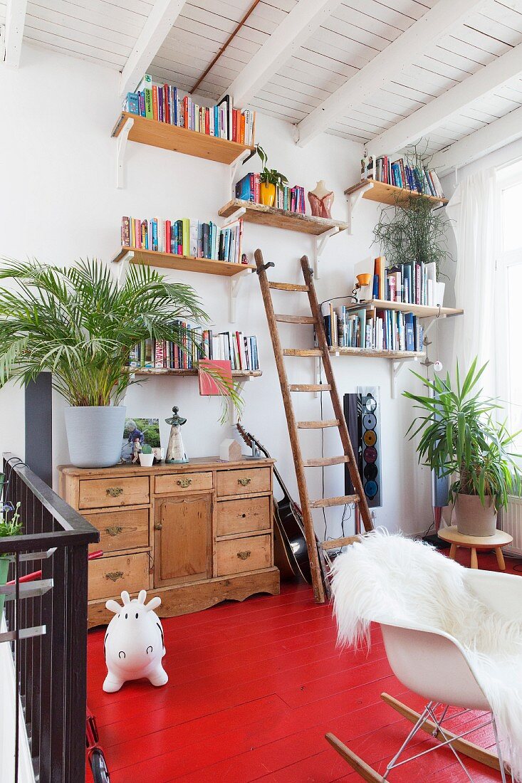 Rot lackierter Holzboden auf Galerie, an der Wand Holzregale mit Büchern, im Vordergrund Klassiker Schaukelstuhl mit weißem Tierfell