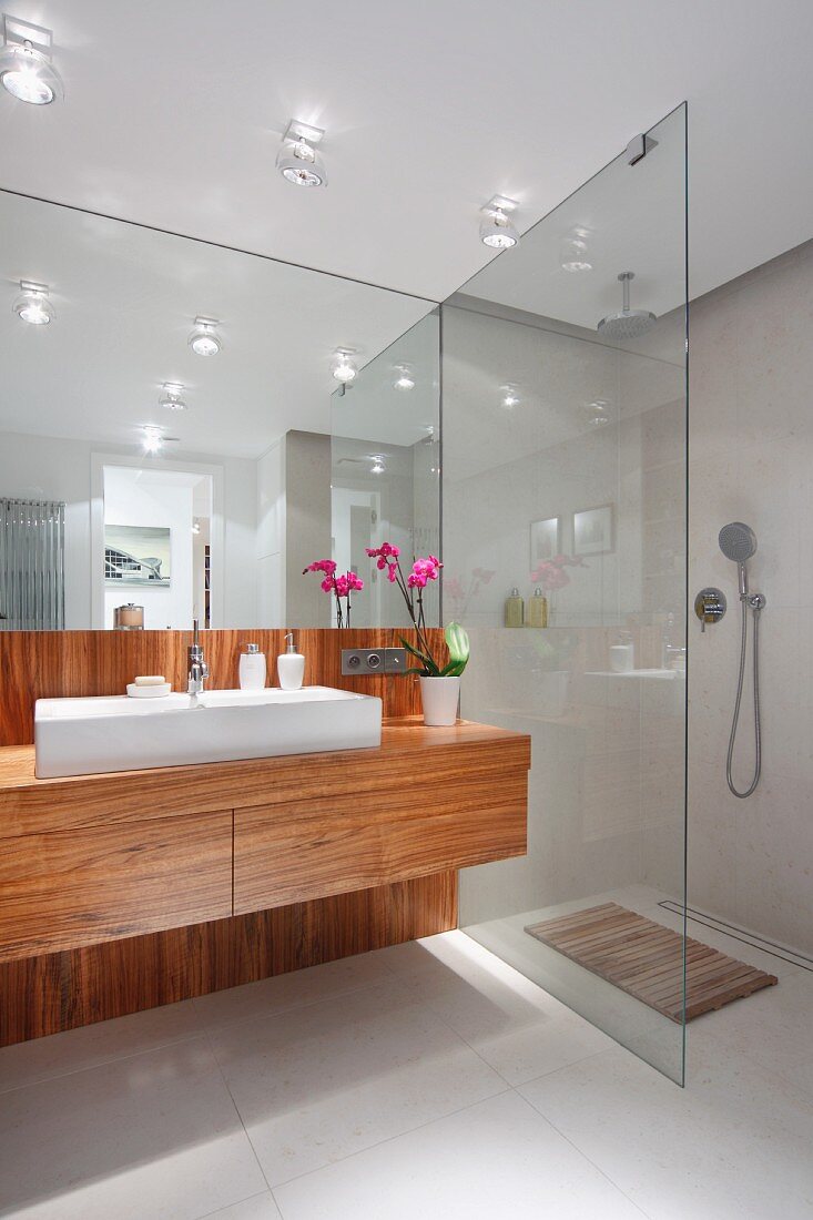 Waschtisch mit trogartigem Waschbecken auf edlem Holzunterschrank und vollflächiger Wandspiegel, seitlich barrierefreie Dusche mit Glas Trennwand