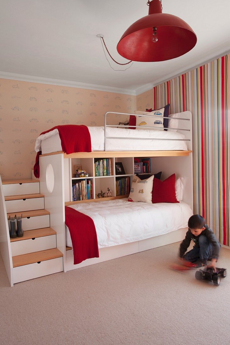 Little boy sitting on floor below pendant lamp with red metal lampshade and bunk beds with stairs in children's bedroom