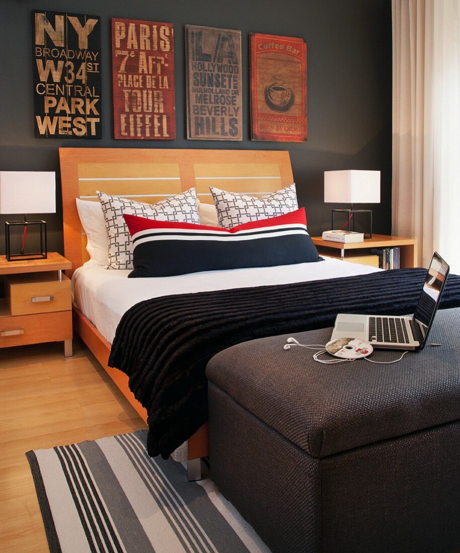 Doppelbett mit Kopfteil aus Holz und rot, weiss, schwarz gestreiftes Kissen, an dunkelgrau getönter Wand alte Ortsschilder, im Vordergrund Laptop auf Polsterhocker