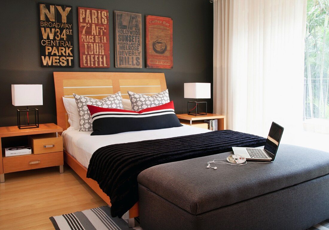 Doppelbett mit Kopfteil aus Holz und rot, weiss, schwarz gestreiftes Kissen, an dunkelgrau getönter Wand alte Ortsschilder, im Vordergrund Laptop auf Polsterhocker, seitlich Fenster mit Vorhang