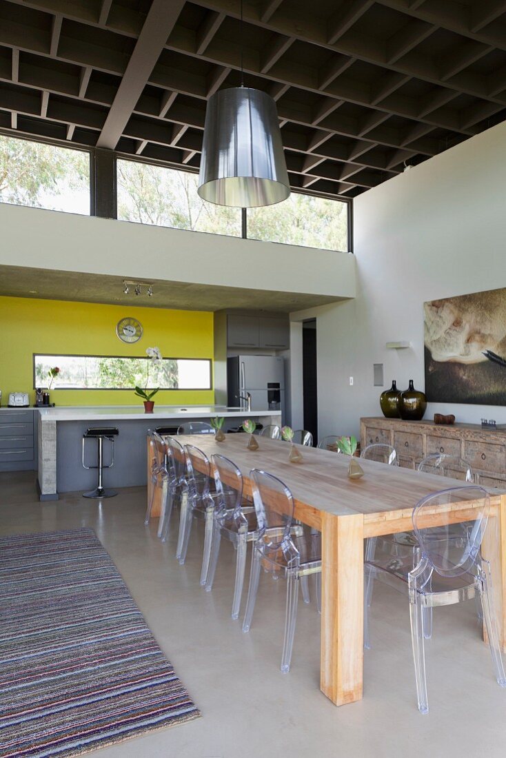 Rustikaler Massivholztisch mit Ghost-Stühlen, im Hintergrund offene Küche mit gelber Wand, kassettenartige Decke