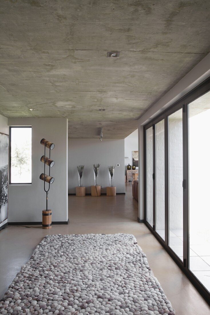 Rug in natural shades in hallway with exposed concrete ceiling, glass wall and plants on tree stump stools in background