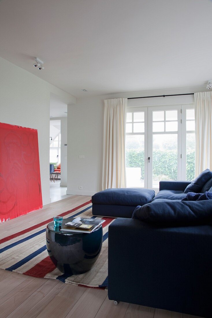 Blaues Sofa und bauchiger Beistelltisch auf gestreiftem Teppich, an Wand rotes Bild gelehnt