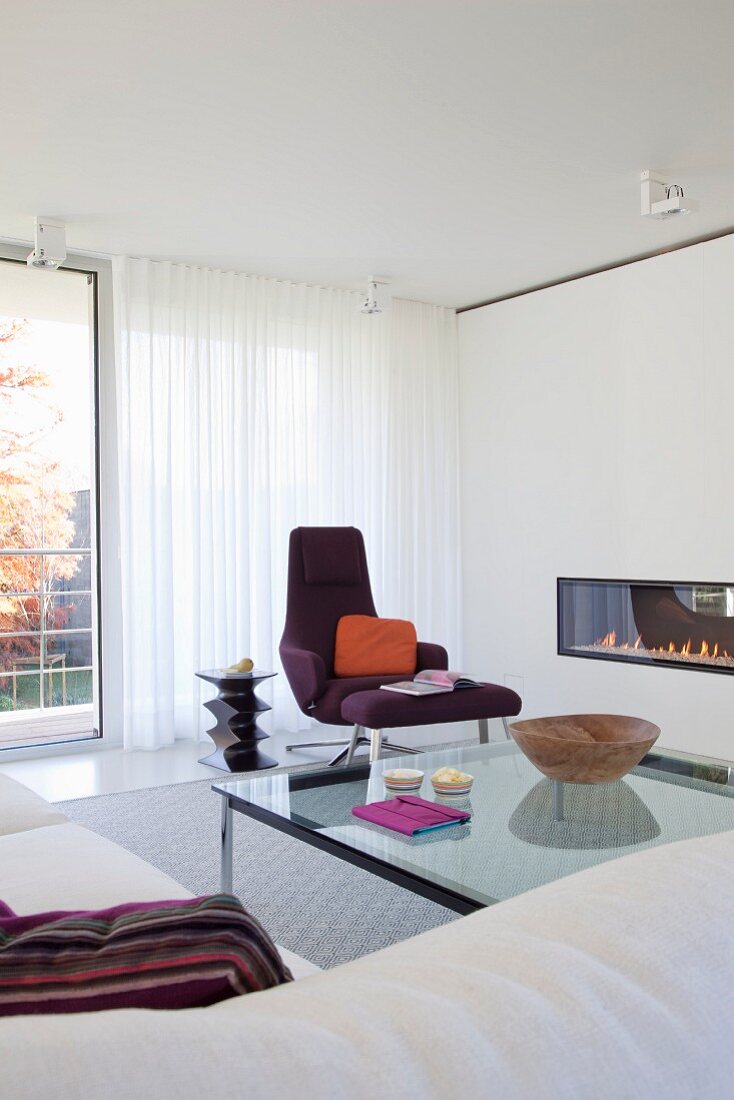 Designermöbel und Feuerstelle in weißer Wandscheibe, bodenlanger Vorhang vor Verglasung in elegantem Wohnzimmer