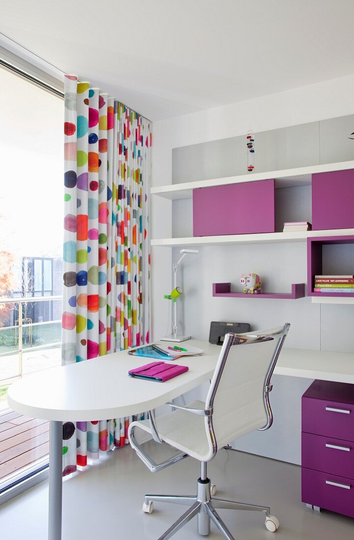 Weißer Schreibtisch und Designer Bürostuhl vor raumhohem Fenster mit bunt gepunktetem bodenlangem Vorhang, seitlich Regalelemente mit violetten Farbakzenten