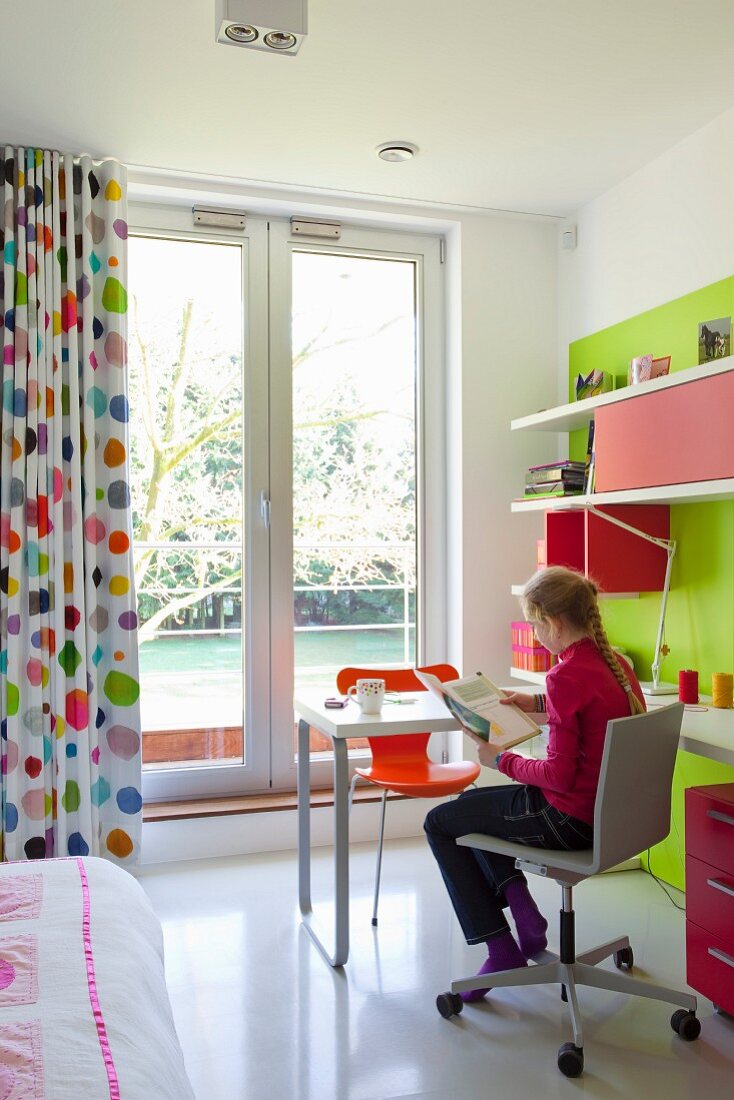 Girl seated at desk in front of French windows with floor-length, multicoloured, polka-dot curtains in child's bedroom