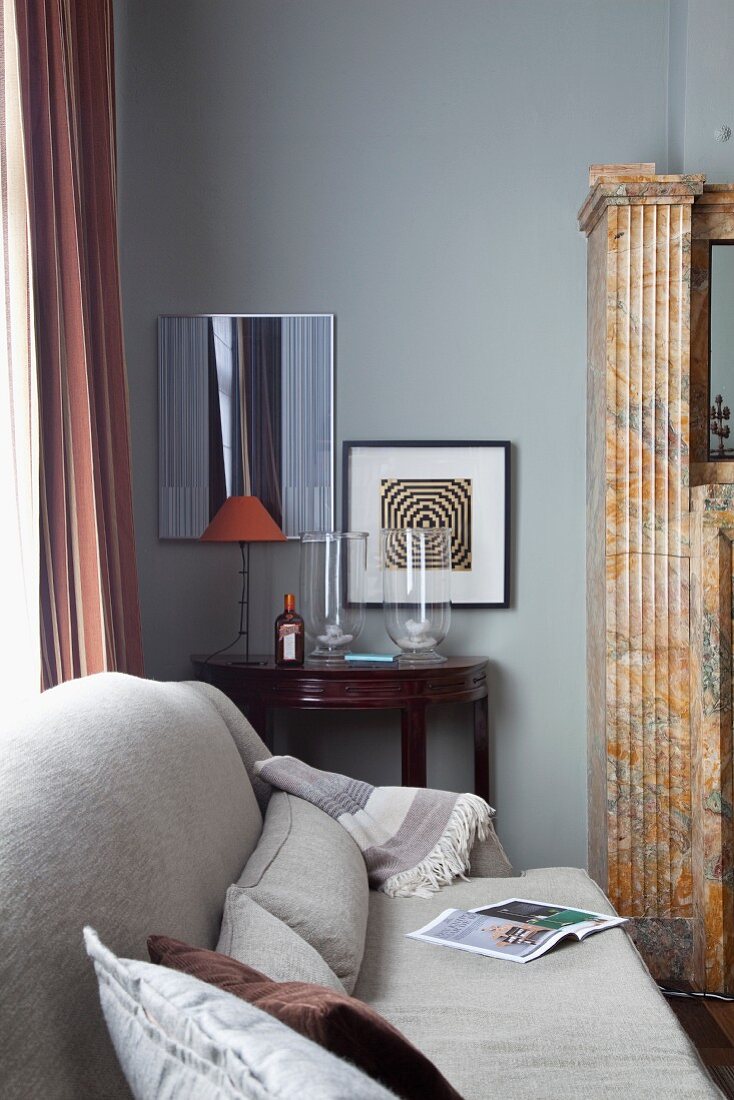 Hellgraue Couch mit Kissen in Wohnzimmerecke, im Hintergrund antiker, halbkreisförmiger Wandtisch an grau getönter Wand