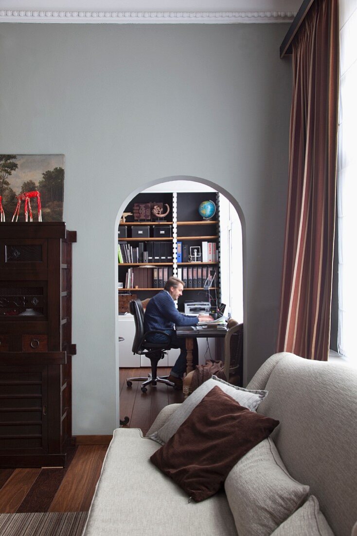 View of man seated at desk seen from living room with pale grey couch through arched doorway