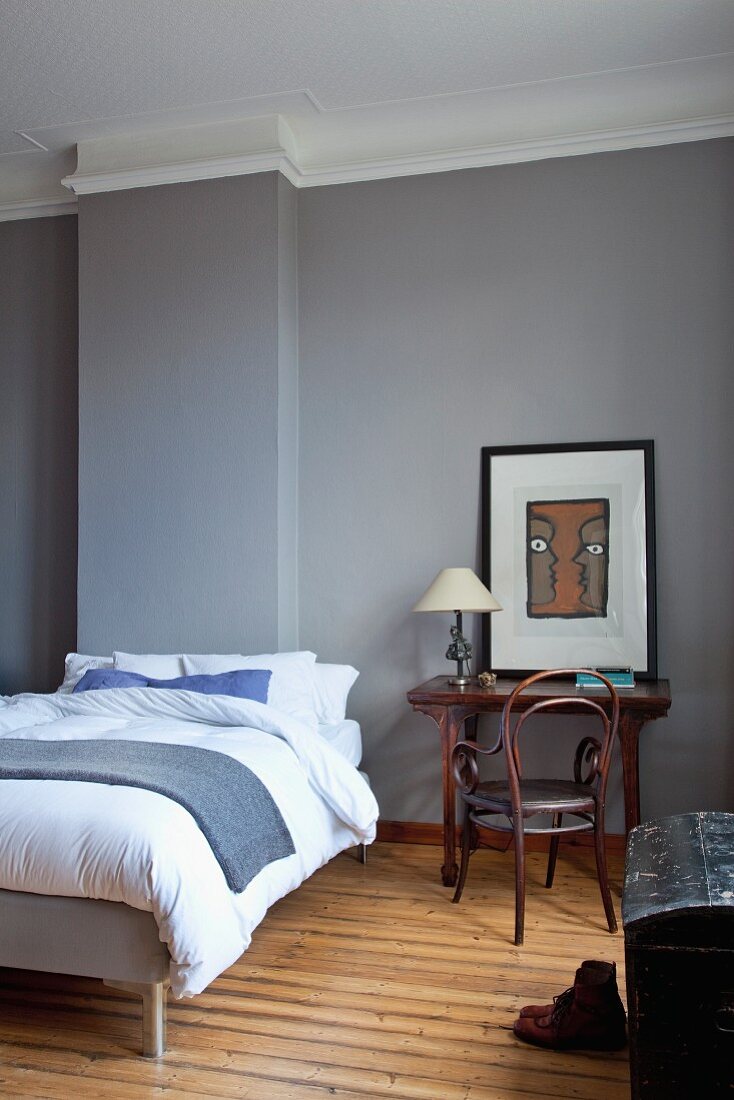 Double bed with pale bed linen next to table and wooden Thonet armchair in grey-painted bedroom