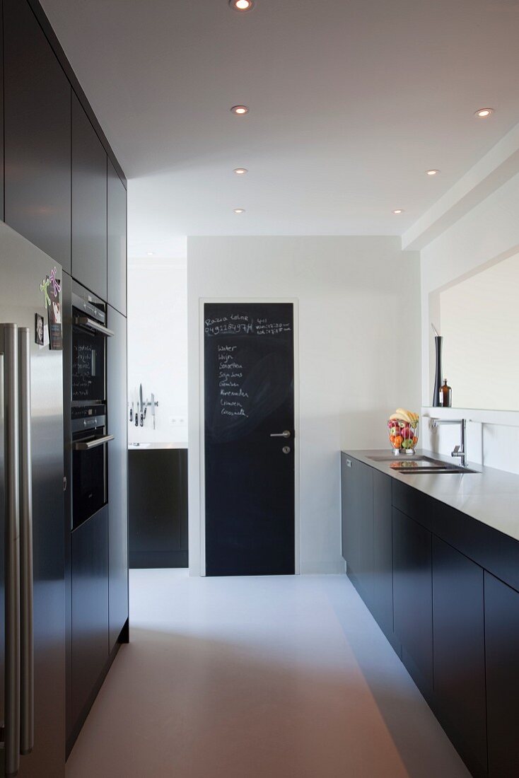 Black designer kitchen with pale worksurface and floor-to-ceiling cupboards with fitted appliances; notes written in chalk on door painted with chalkboard paint