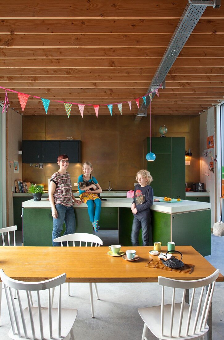 Offene Wohnküche mit Mutter und zwei Kindern an Kochinsel; vorne Esstisch mit Stühlen