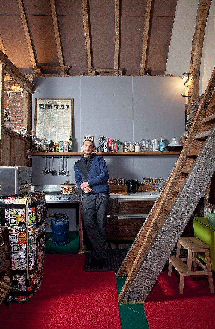 Young man in DIY loft-apartment kitchen with rustic ladder leading to gallery and retro fridge covered in colourful stickers on red carpet