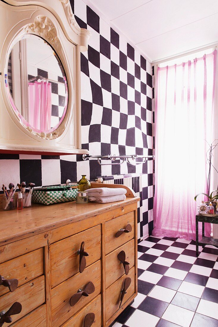 Schachbrettmuster auf Boden und an Wand, rosa Vorhang am Fenster, im Vordergrund rustikaler Schubladenschrank aus Holz