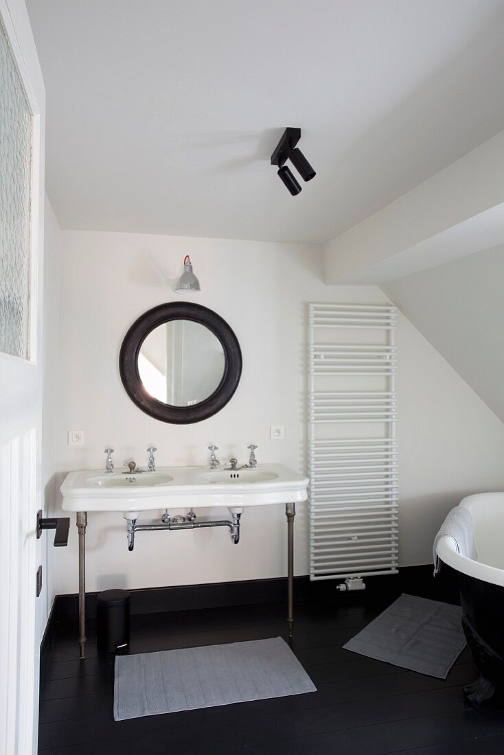 Retro Waschtisch mit Metallfüssen unter rundem Spiegel mit schwarzem Rahmen, seitlich Handtuchtrockner an Wand