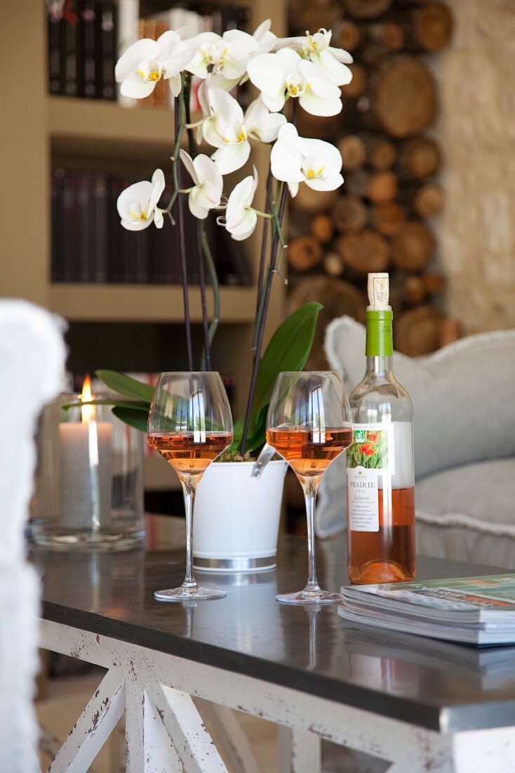 Potted white orchid, bottle and two glasses of rose wine on coffee table in elegant, country-house interior