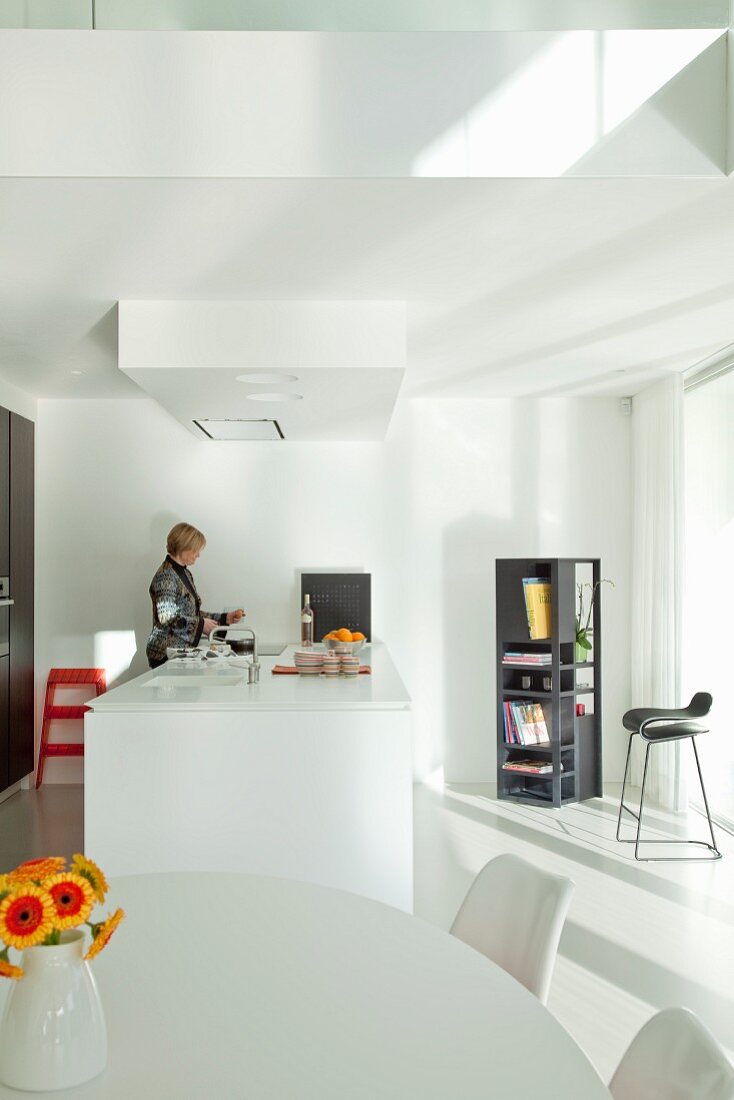 Open-plan, white designer kitchen with dining area and kitchen counter; woman in background