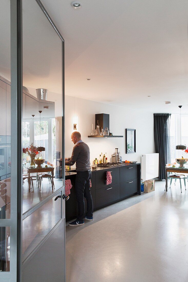 Blick durch offene Tür in Küche mit schwarzen Unterschränken und Betonboden, Mann vor Küchenzeile