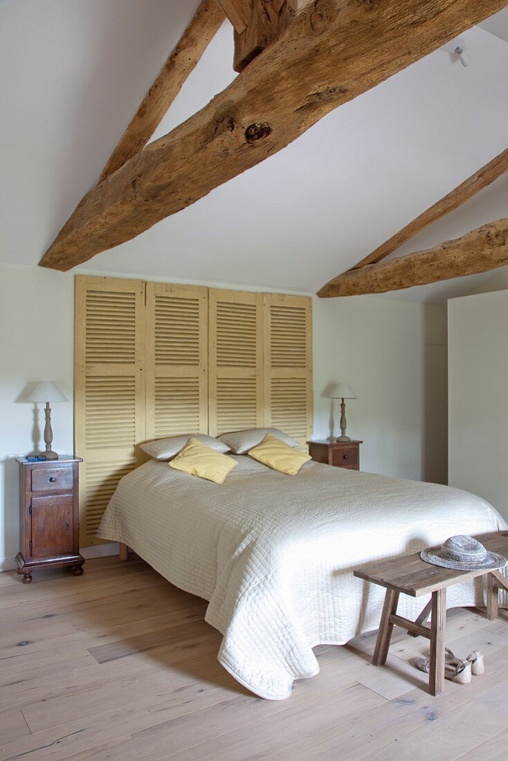 Double bed with white bedspread in front of closed shutters in renovated attic bedroom with exposed wooden roof structure