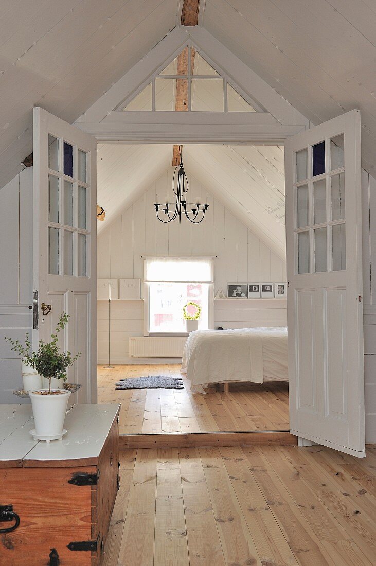 Blick durch geöffnete Sprossen-Flügeltür in einfaches weißes Landhausschlafzimmer im Dachgeschoss mit naturbelassenem Dielenboden und skandinavischem Vintage Flair
