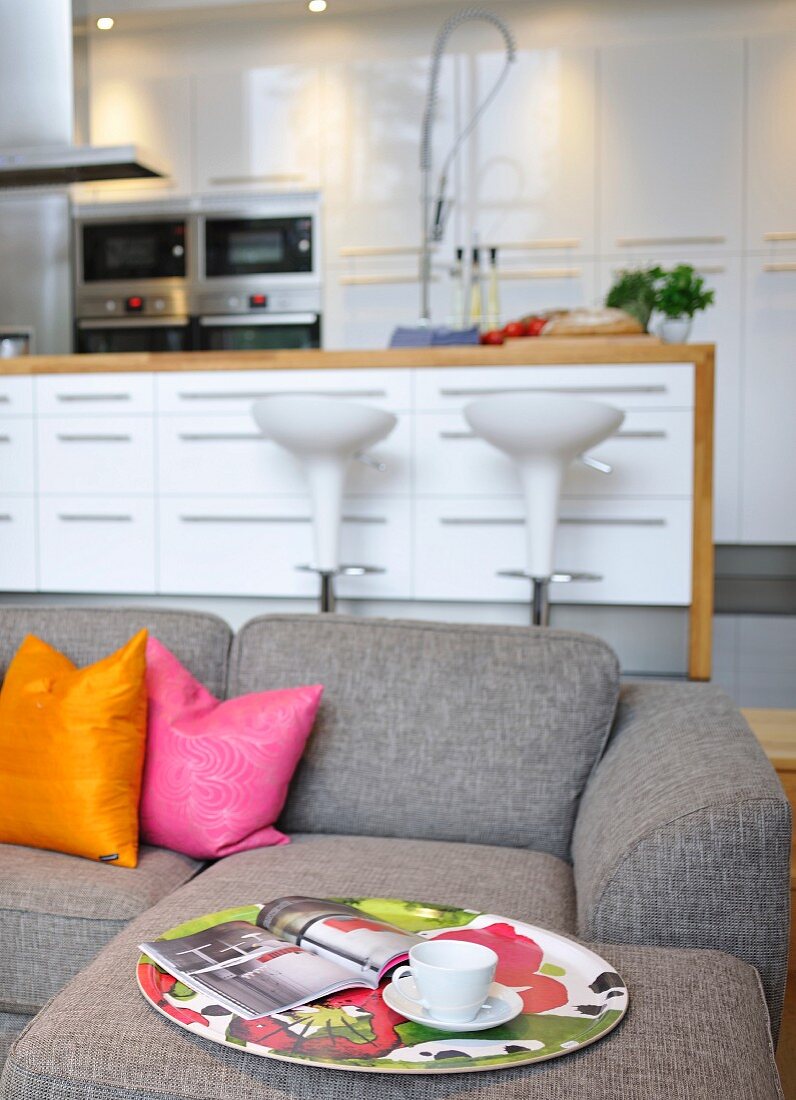 Graues Polstersofa mit farbigen Kissen; im Hintergrund Barhocker vor Kücheninsel