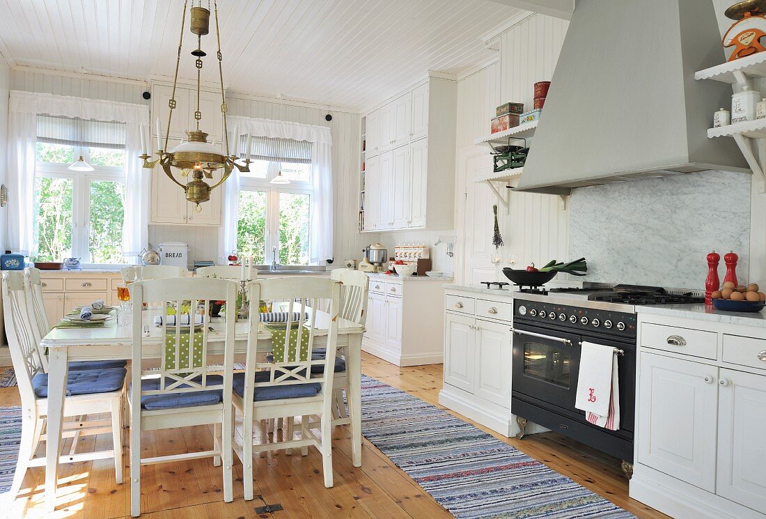 Square dining table and chairs below brass lamp and woven rugs in spacious, white, Scandinavian, country-house kitchen