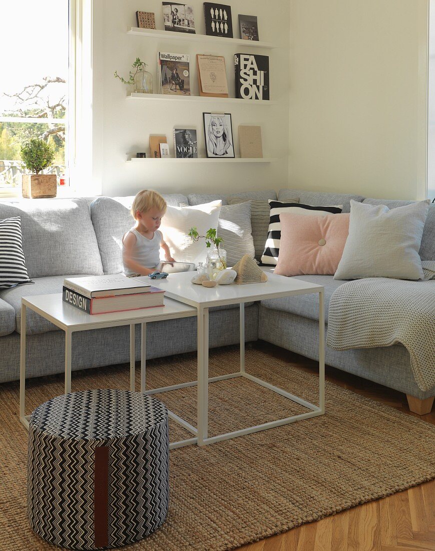 Gemütliches Wohnzimmer mit Kleinkind auf grauem Polstersofa und Wandboards mit Büchern
