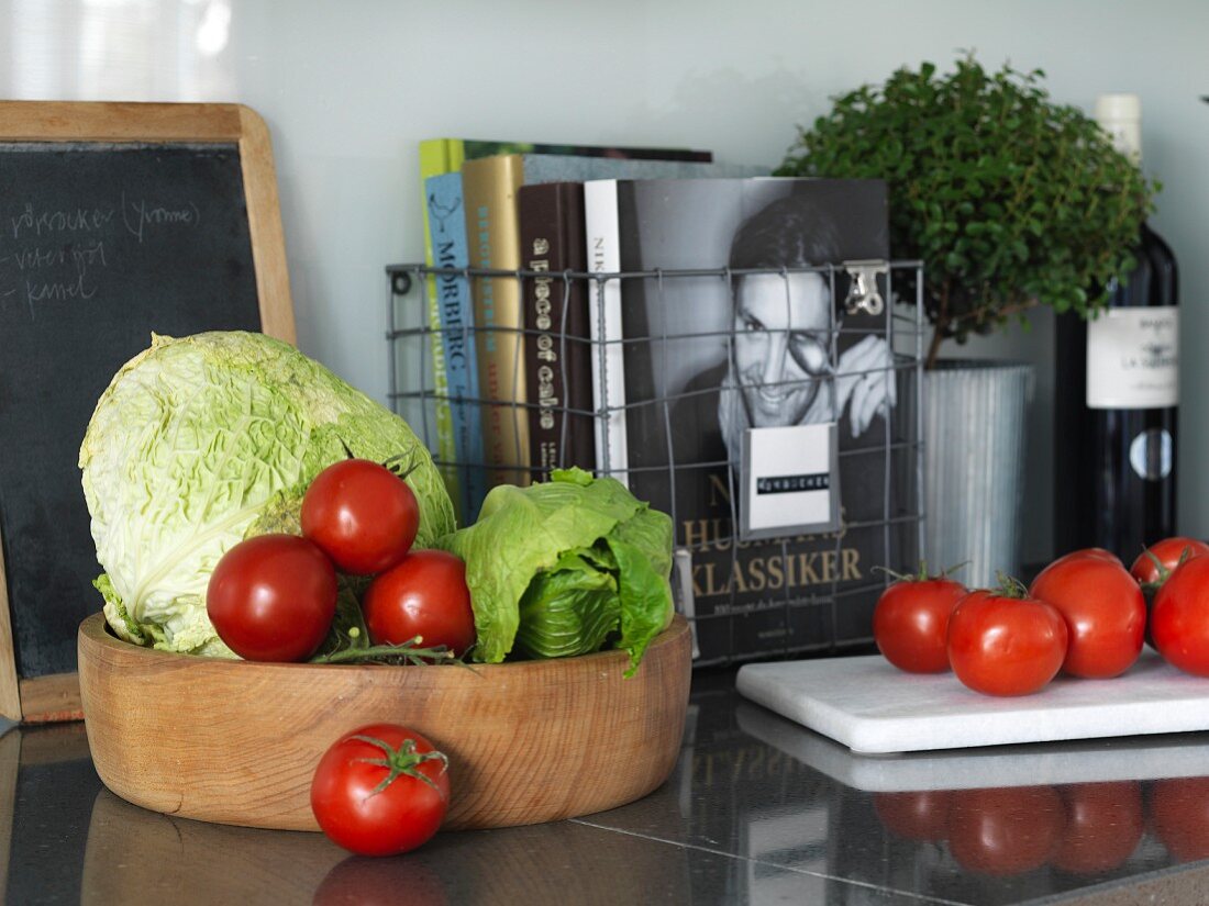 Drahtkorb mit Büchern auf Granitküchenarbeitsplatte und Holzschale mit Gemüse vor nostalgischer Schiefertafel