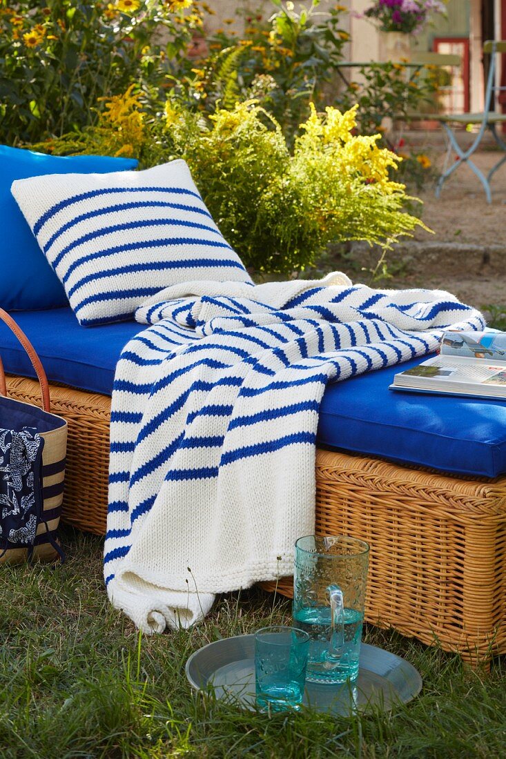 A wicker lounger with a blue cushion in a garden with a stripped throw and a cushion