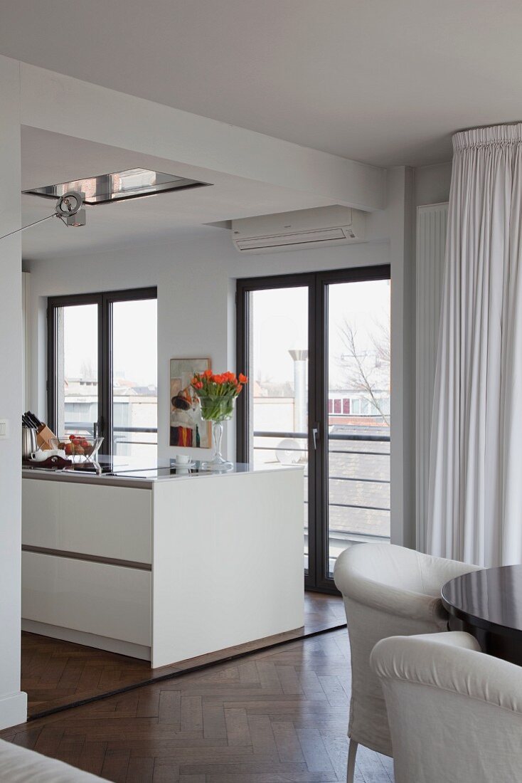 Teilweise sichtbarer Essplatz mit weissen Sesseln, im Hintergrund weiße Kücheninsel vor Balkontür