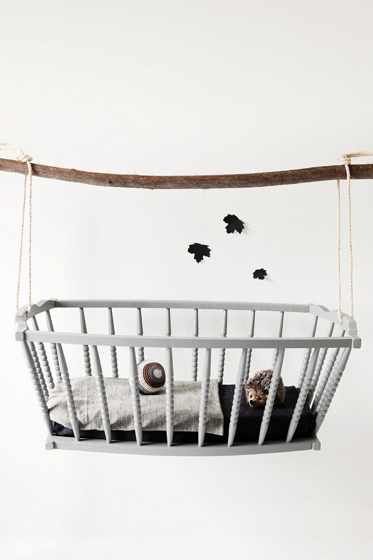 An Holzstock aufgehängte Puppenwiege mit Igelfigur und Strickball; aus schwarzem Papier ausgeschnittene Blätter an der Wand
