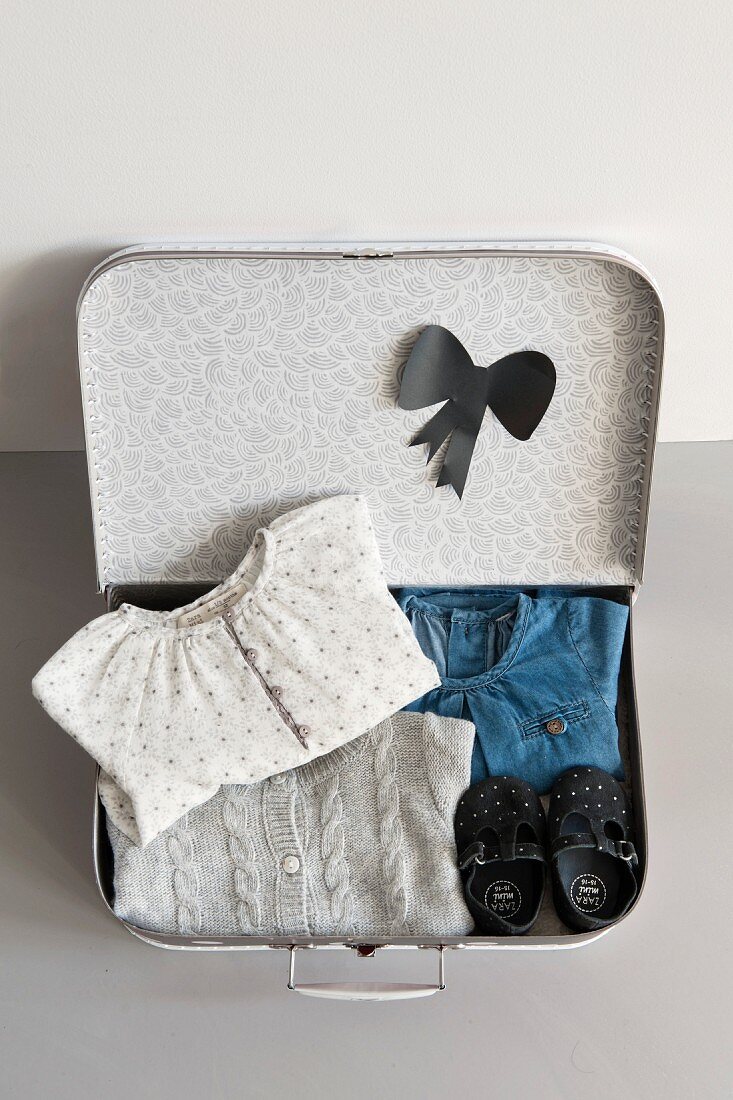 Aufgeklappter Kinderkoffer mit zusammengelegter Mädchenkleidung und aus schwarzem Papier ausgeschnittener Kragenschleife