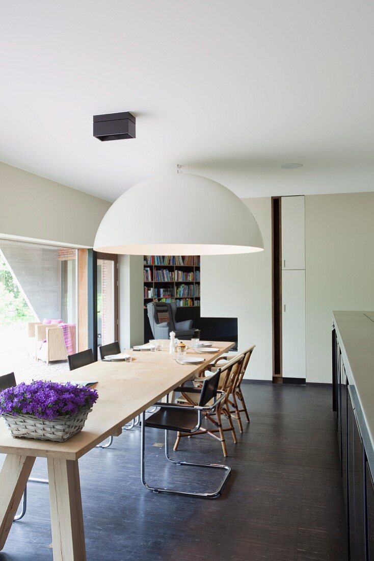 Lange Tafel aus hellem Holz mit klassischen Freischwingern und riesiger Halbkugelleuchte an der Decke in offener Küche
