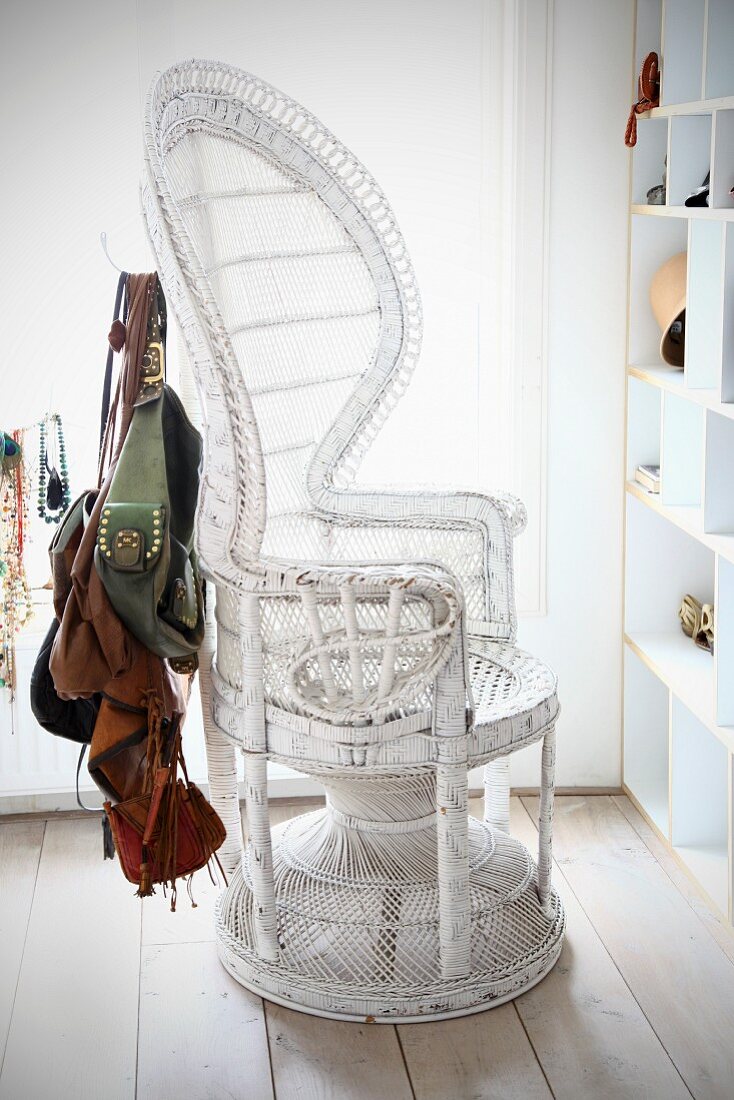 Collection of handbags hanging from white, wicker empress chair next to open-fronted shelving