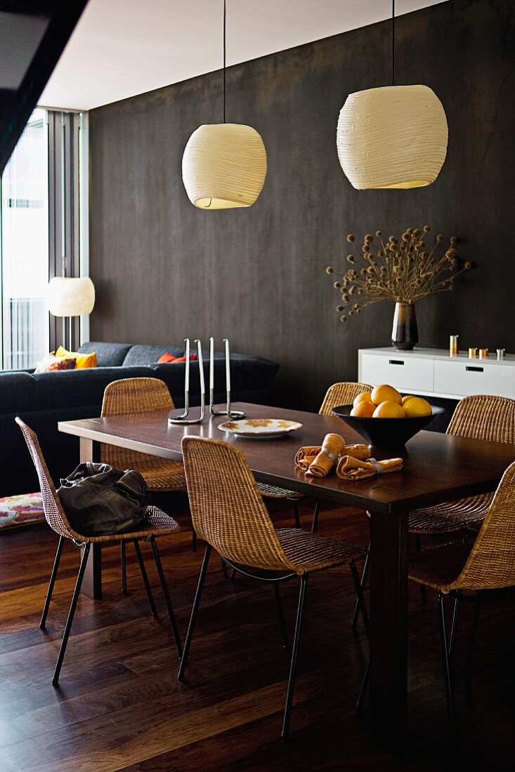 Dark wooden dining table, 60s wicker chairs and pale pendant lamps in front of black wall