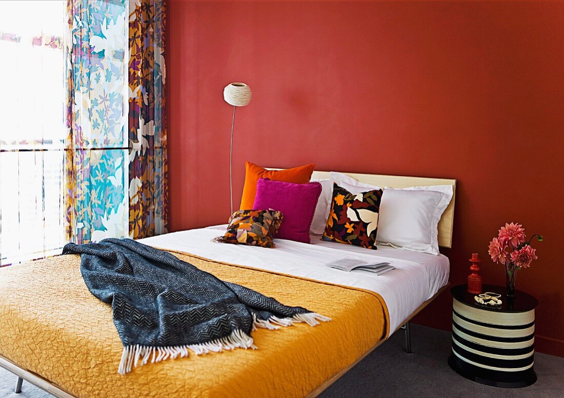 Double bed with yellow bedspread and colourful scatter cushions and striped bedside table against terracotta-red wall