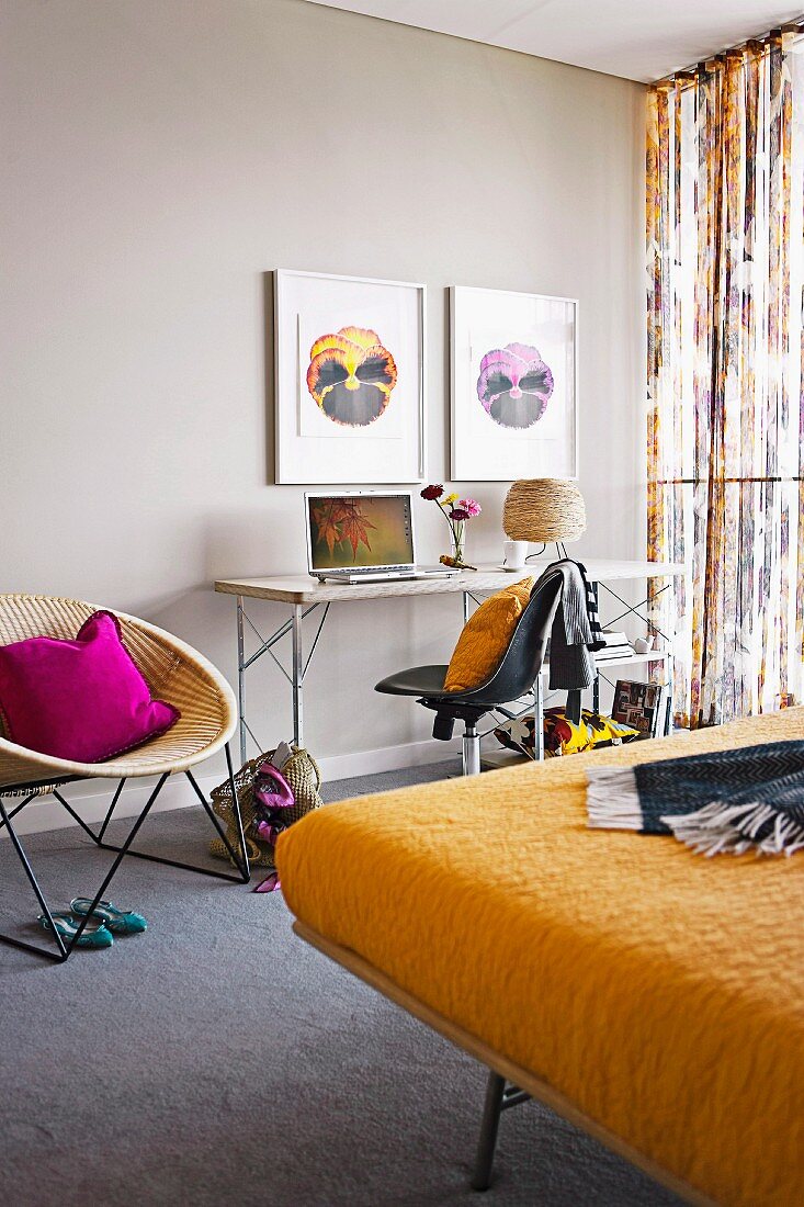 Bilder mit Stiefmütterchenblüten über schmalem Schreibtisch und 60er Jahre Korbstuhl, Doppelbett mit gelber Decke im Schlafzimmer