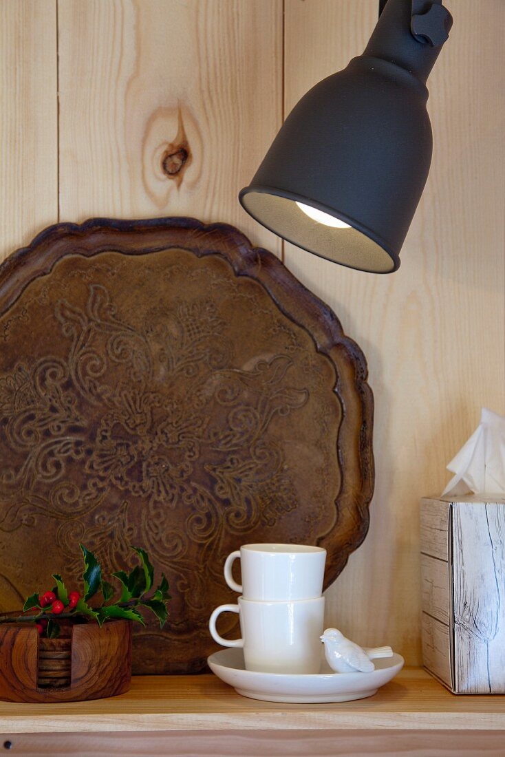 White espresso cups and vintage tray on shelf below wall lamp on wooden wall