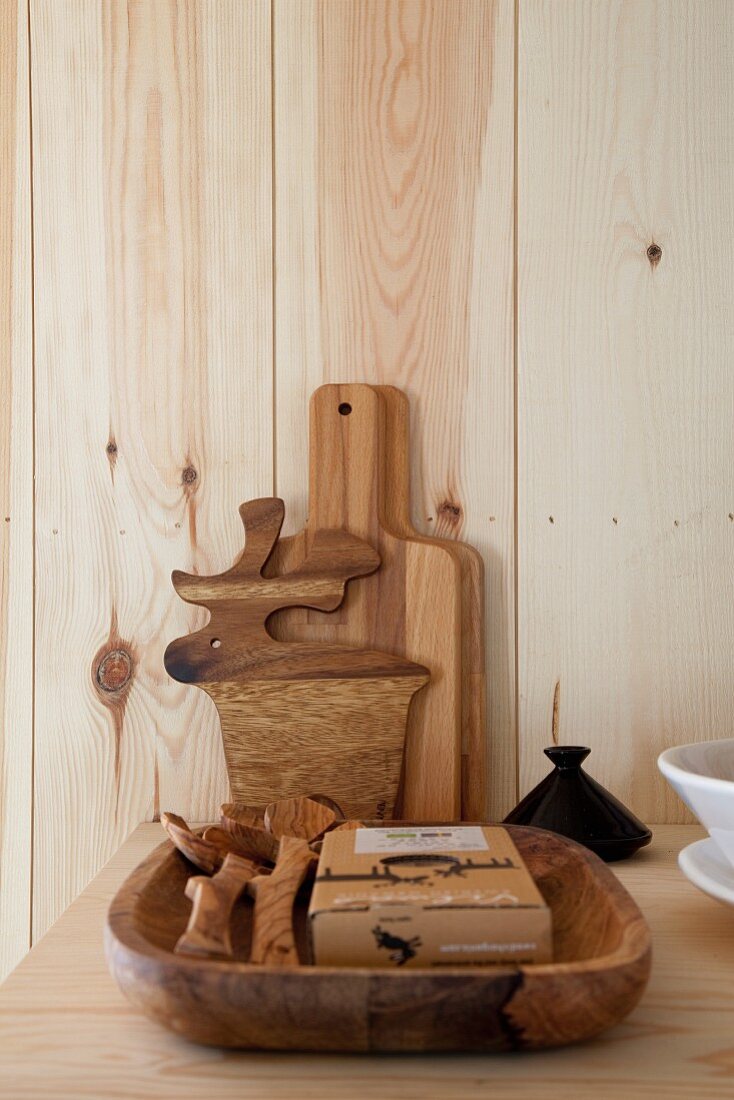 Solid wooden dish of kitchen utensils and chopping boards leaning against wooden wall