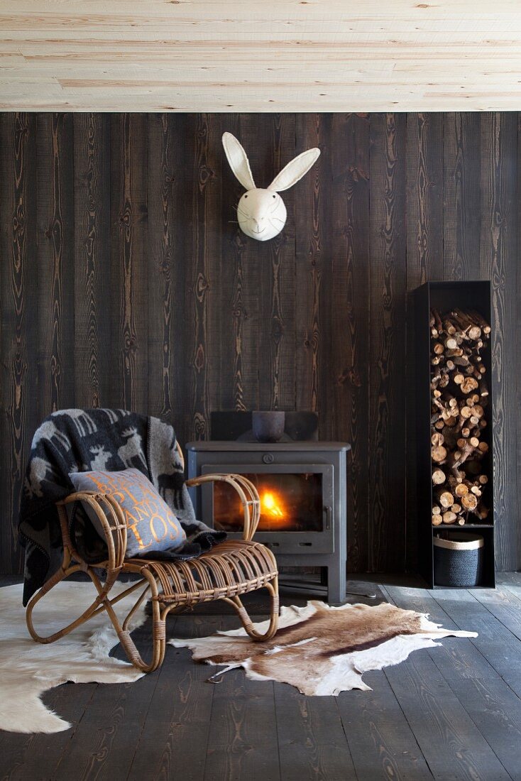 Wicker armchair on animal-skin rugs in front of wood-burning stove below stylised bunny head on dark brown wooden wall