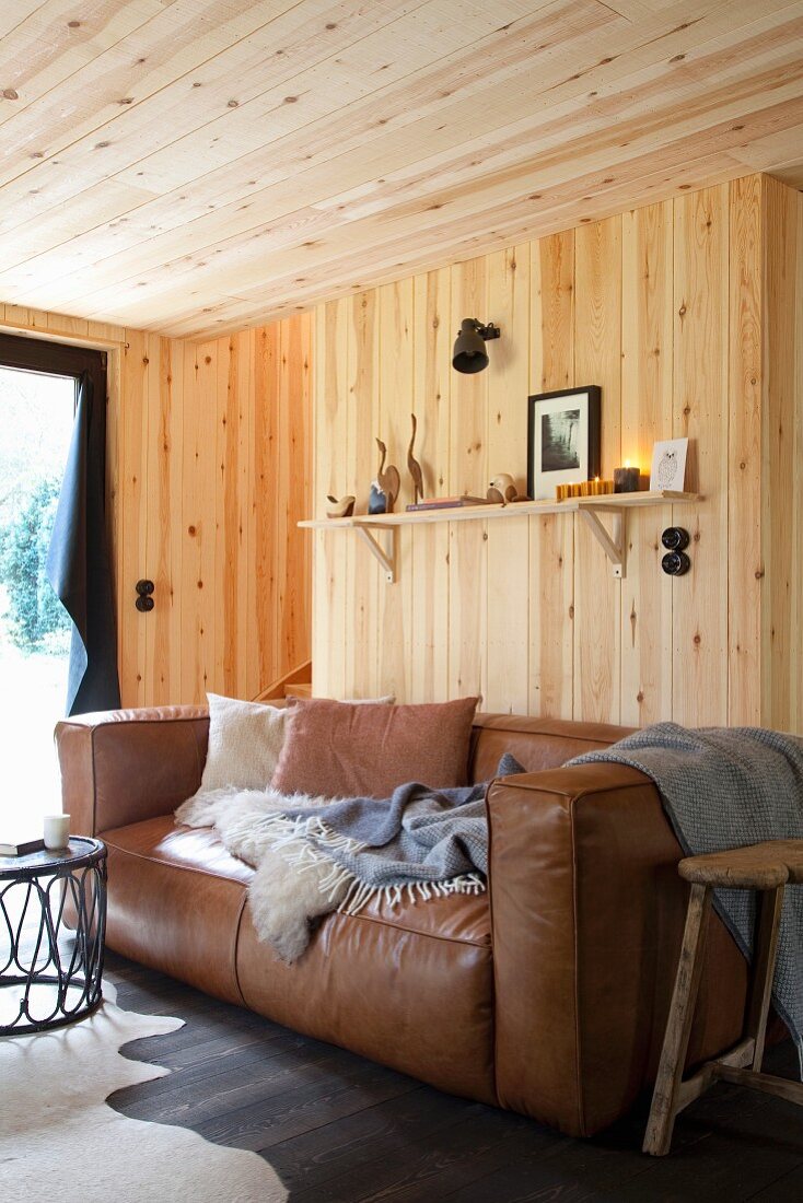 Gemütliche hellbraune Ledercouch mit Kissen, Schaffell und Plaid in holzverkleidetem Wohnraum