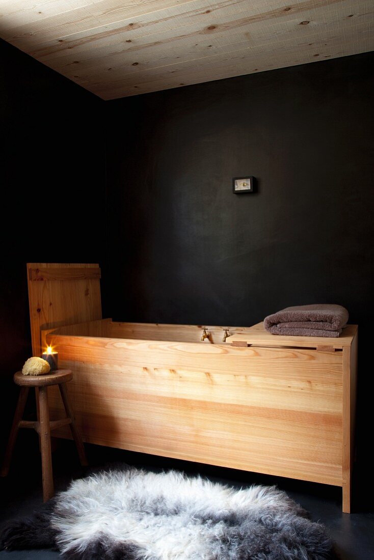 Holz-Badewannentrog vor schwarzer Wand mit schwarz-weißem Schaffell auf schwarzen Boden und Kerzenlicht