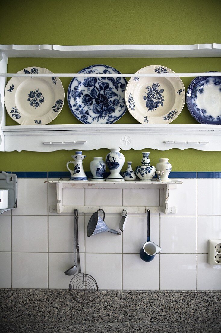 Kleines Wandbord mit Küchenutensilien an gefliester Wand, unter geschnitztem Bord mit weiss-blau bemalten Tellern