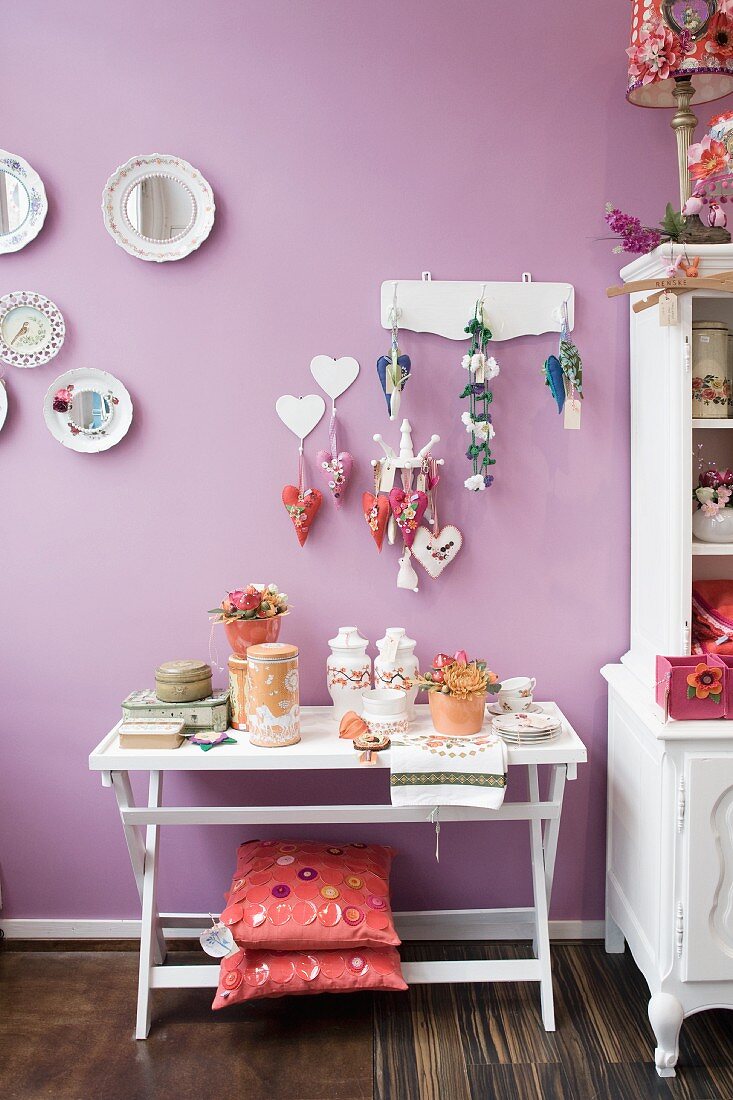 Ornaments on white console table with cushions on base frame below decorations hanging from hooks and painted plates on lilac wall
