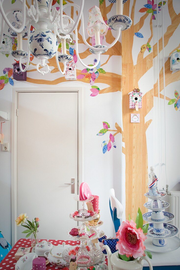 Mehrarmiger Kronleuchter über dekoriertem Tisch mit romantischem Charme, an Wand gemaltes Baummotiv und aufgehängte Vogelhäuschen