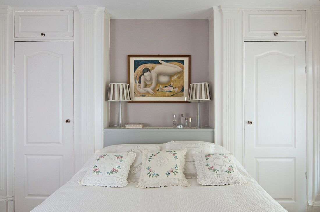 Bed with floral, lace scatter cushions below painting of woman in niche flanked by fitted wardrobes with panelled doors