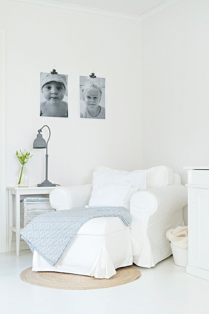 Weisser Polstersessel in Zimmerecke, graue Lampe auf Beistelltisch und schwarz-weiße Fotos an der Wand