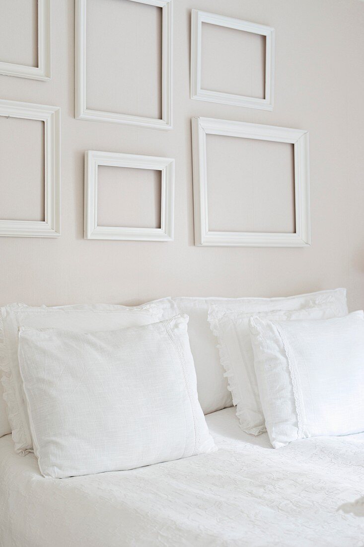 Double bed with white bed linen and stacked pillows below empty, white picture frames on wall